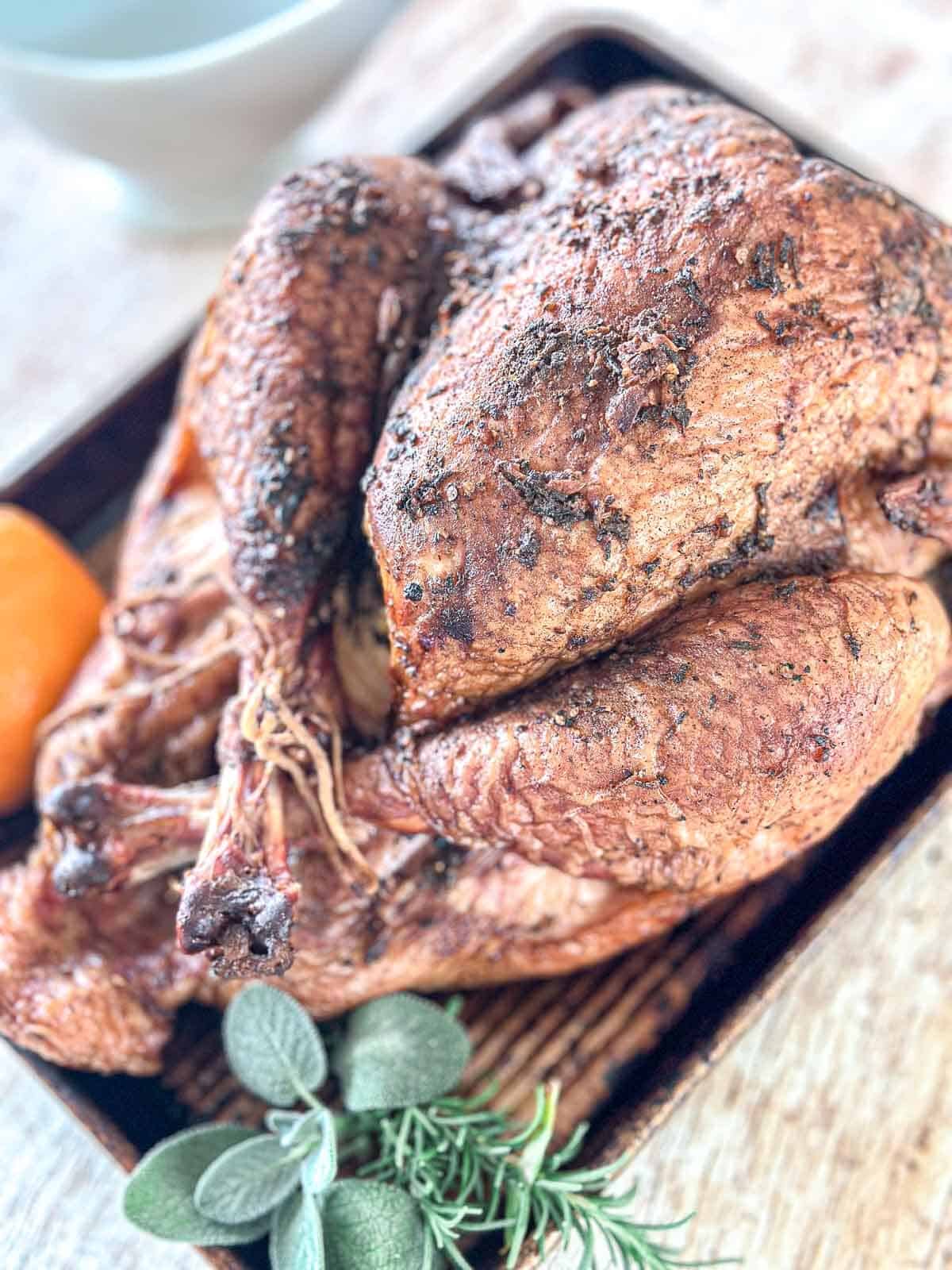 A smoked whole turkey seasoned with herbs sits in a pan, garnished with fresh rosemary and sage; an orange rests nearby on a wooden surface.