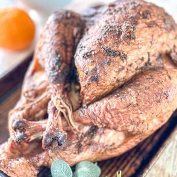 A close-up of a whole roasted turkey with crispy, seasoned skin on a wooden board. A blurred orange and some fresh sage leaves are visible in the background.