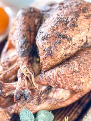 A close-up of a whole roasted turkey with crispy, seasoned skin on a wooden board. A blurred orange and some fresh sage leaves are visible in the background.