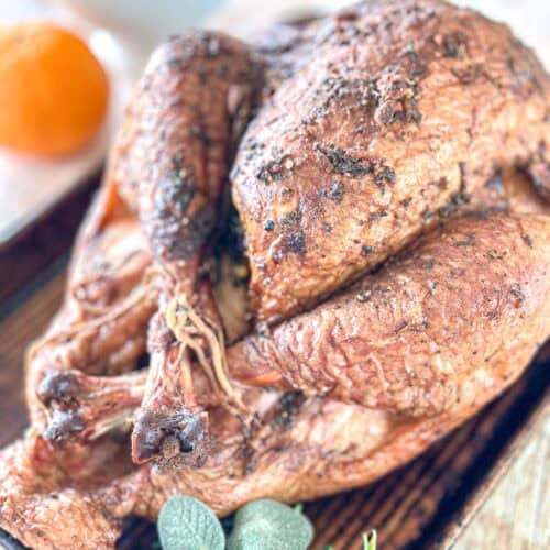 A close-up of a whole roasted turkey with crispy, seasoned skin on a wooden board. A blurred orange and some fresh sage leaves are visible in the background.
