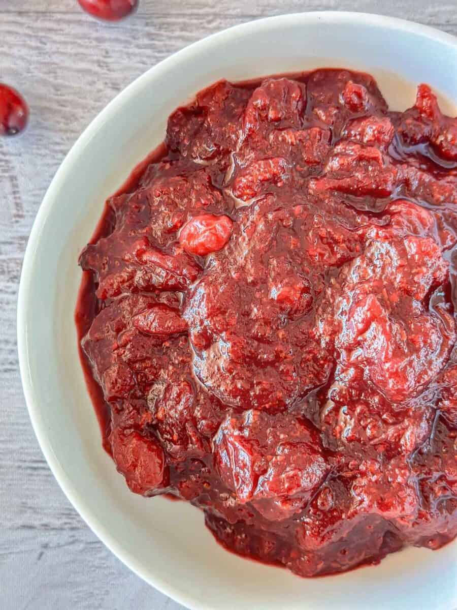 A close-up of a white bowl filled with thick, chunky cranberry sauce on a light gray surface. A few whole cranberries are visible in the background.