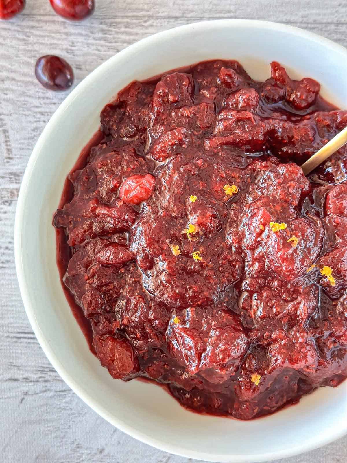 A white bowl filled with chunky cranberry sauce sits on a light gray surface. A few whole cranberries are nearby, and small bits of orange zest are visible on top of the sauce.