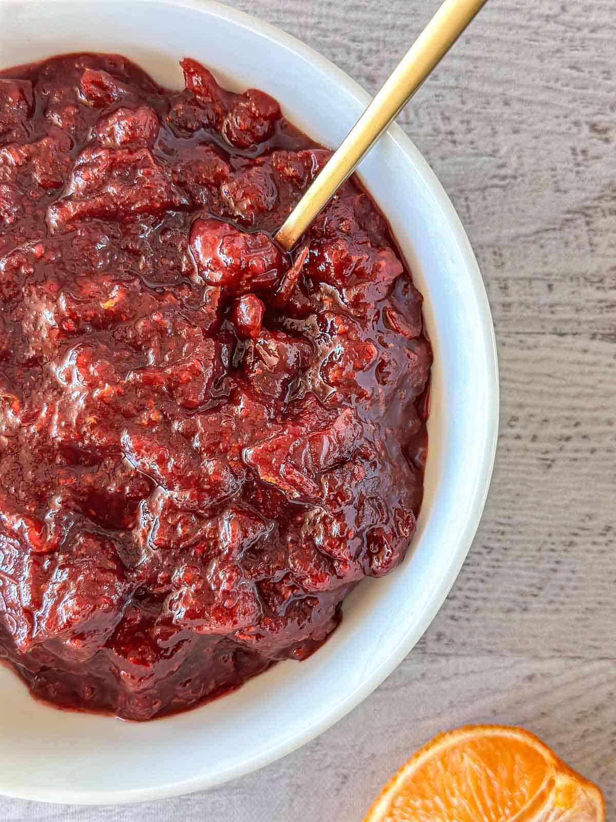 A white bowl filled with chunky cranberry sauce, with a gold spoon resting inside. The bowl is placed on a light gray surface, and an orange slice is partially visible in the bottom right corner.