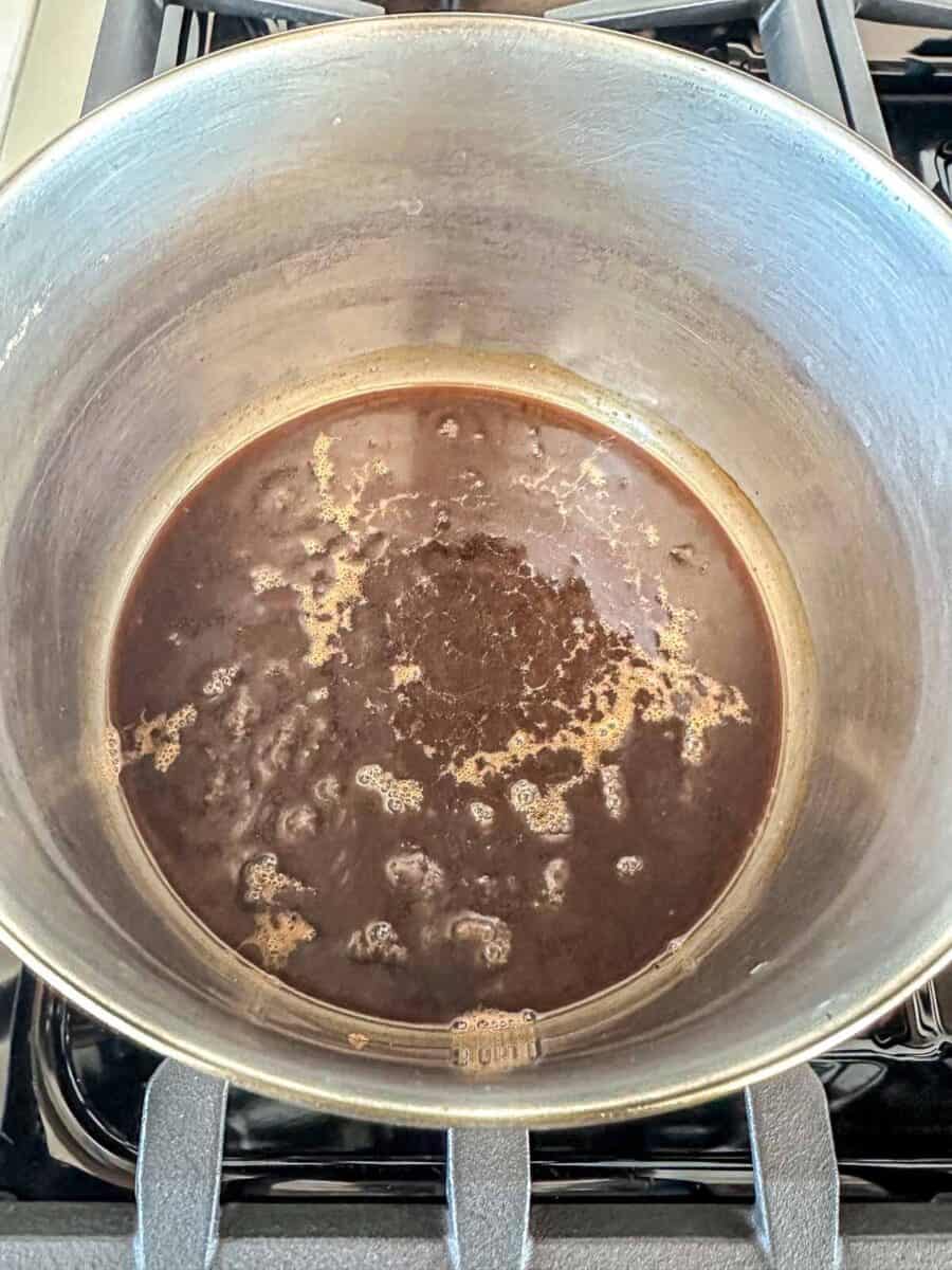A large metal pot sits on a stovetop, containing a dark brown liquid that appears to be simmering, with bubbles and foam forming on the surface.