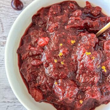 A close-up of a bowl filled with thick, chunky homemade cranberry sauce, garnished with small bits of orange zest, with a spoon partially visible on the right side.