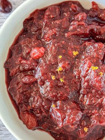 A close-up of a bowl filled with thick, chunky homemade cranberry sauce, garnished with small bits of orange zest, with a spoon partially visible on the right side.