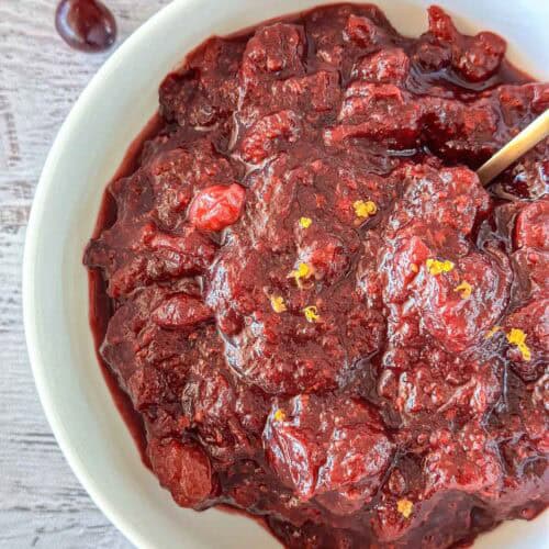 A close-up of a bowl filled with thick, chunky homemade cranberry sauce, garnished with small bits of orange zest, with a spoon partially visible on the right side.