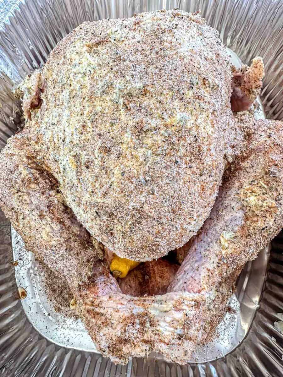 A raw turkey coated in a dry seasoning rub sits in a large metal roasting pan, with a piece of lemon visible inside the cavity.