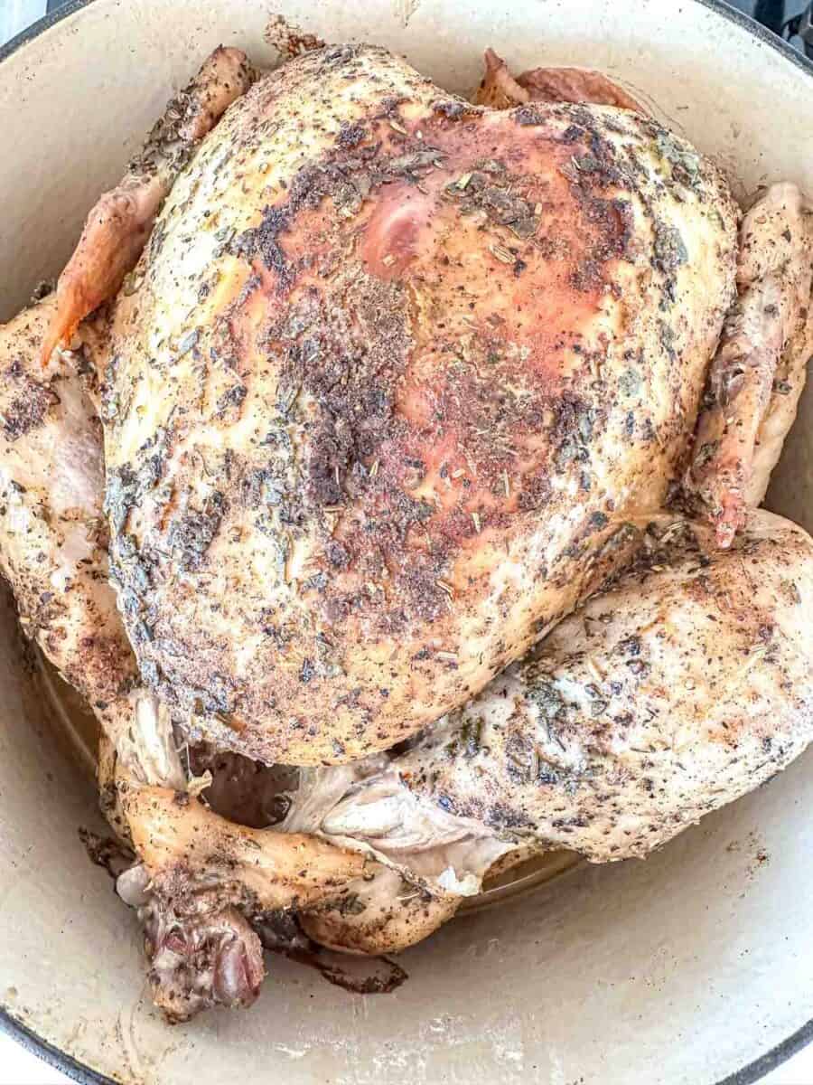 A whole roasted turkey seasoned with herbs and spices sits in a large white pot, showing golden-brown, crispy skin.