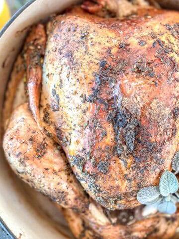 A whole roasted turkey with crispy, seasoned skin sits in a large pot, garnished with fresh sage leaves. The chicken is golden brown and surrounded by herbs.