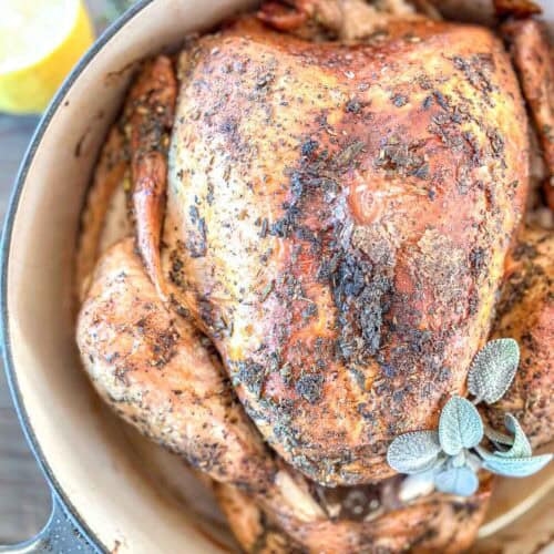 A whole roasted turkey with crispy, seasoned skin sits in a large pot, garnished with fresh sage leaves. The chicken is golden brown and surrounded by herbs.