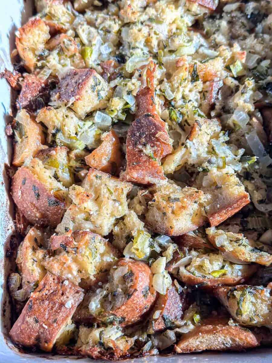 A close-up of baked sourdough bread stuffing with golden-brown bread cubes, chopped celery, onions, and herbs in a white baking dish.
