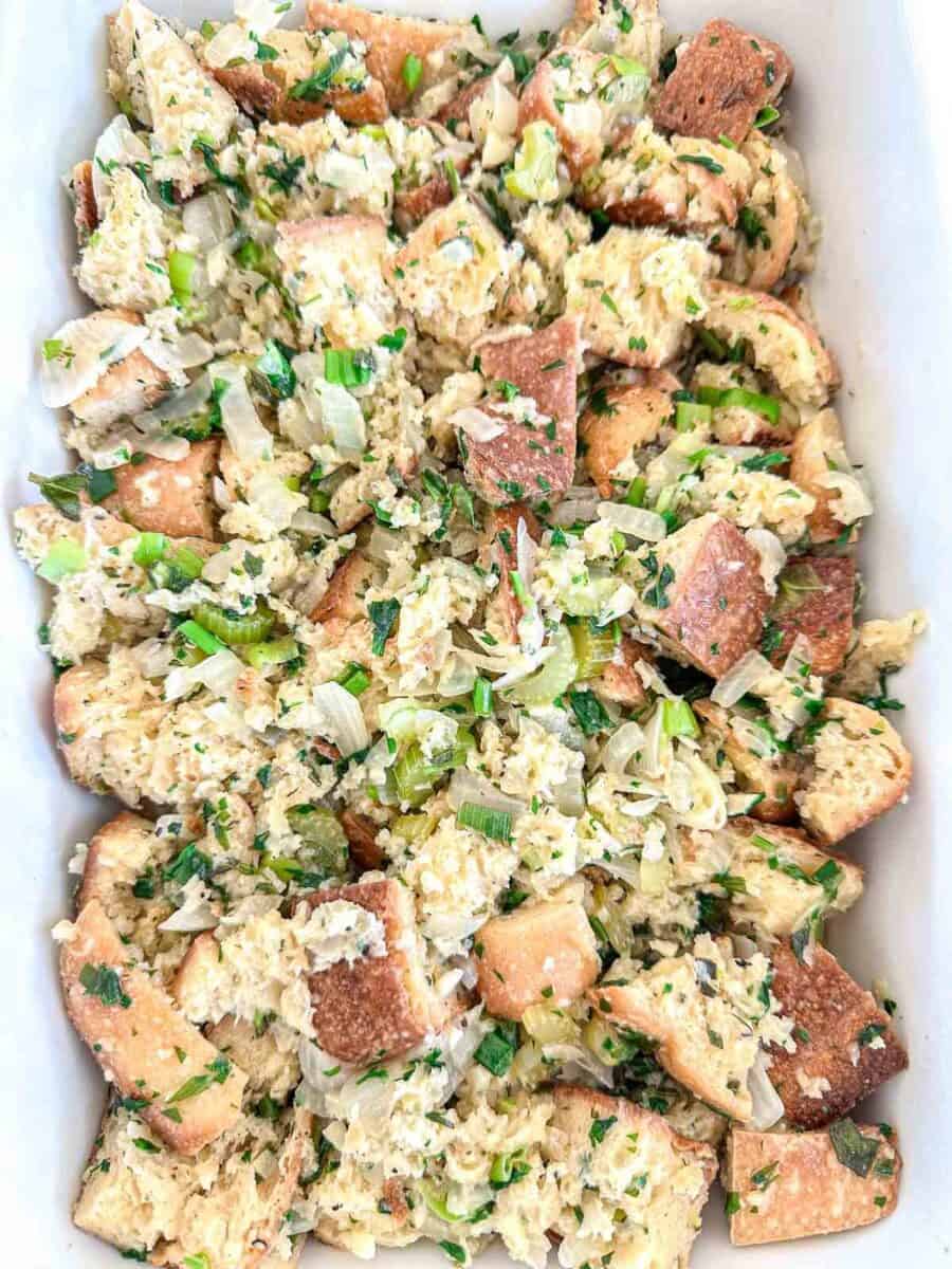 A close-up of a baking dish filled with chunky bread stuffing mixed with herbs, chopped celery, and onions, ready to be baked.