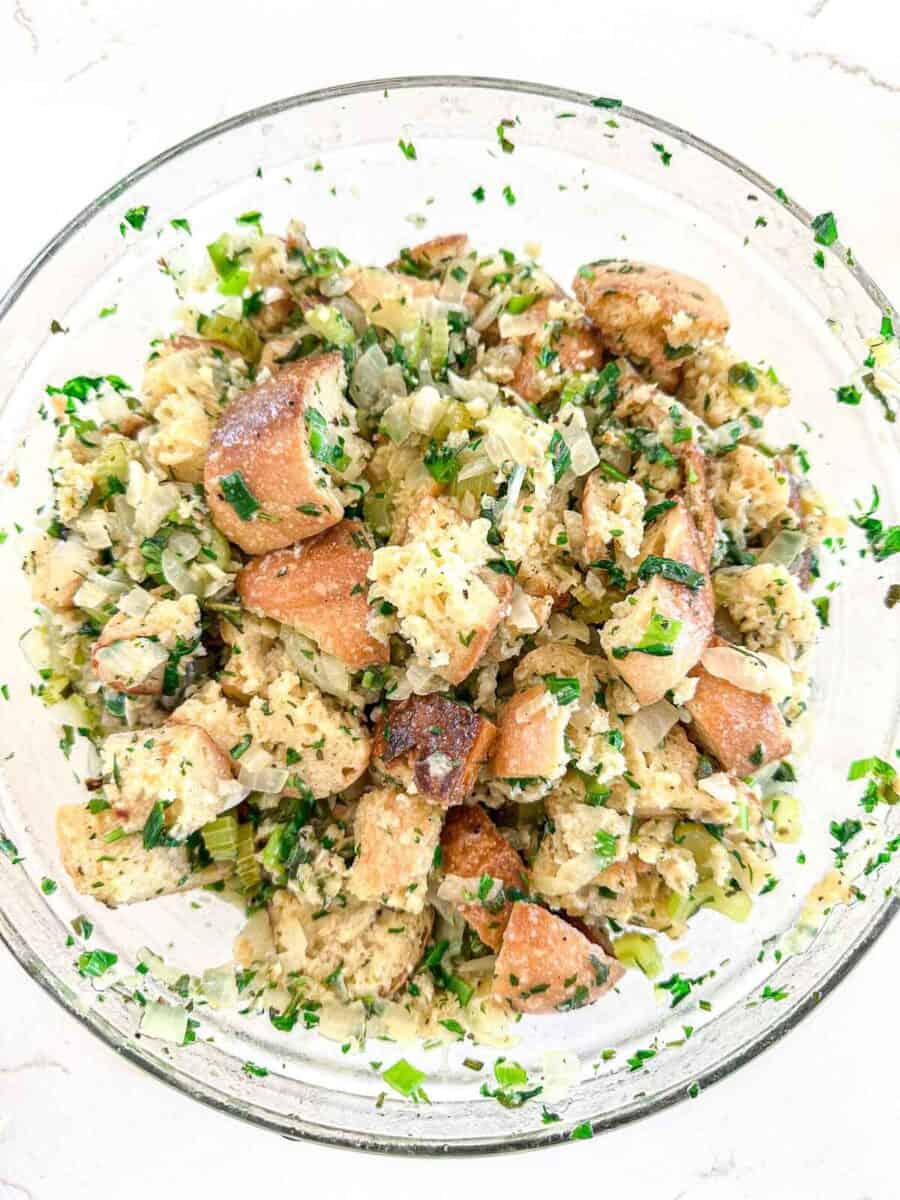 A glass bowl filled with homemade stuffing made of bread cubes, chopped herbs, onions, and celery, all mixed together. The stuffing appears fresh and lightly toasted with green and brown tones.