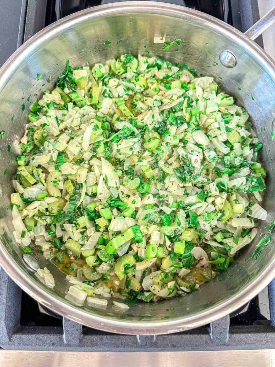 Chopped onions, celery, and parsley are sautéing in a stainless steel pan on a stovetop. The vegetables are lightly cooked and mixed with herbs.