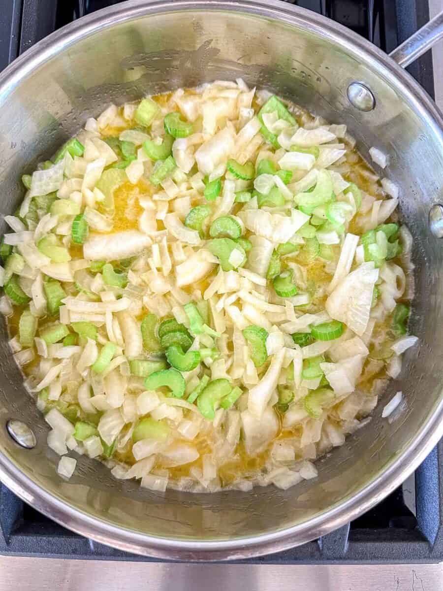Chopped onions and celery sautéing in melted butter in a stainless steel pan on a stove.
