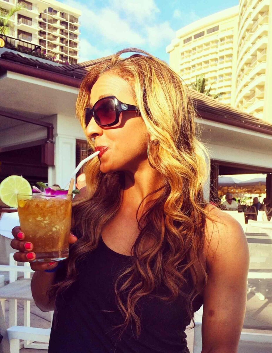 Woman with long wavy hair and sunglasses sips a tropical drink with a straw. The drink is garnished with a lime and flower. She is outdoors at a sunny resort with tall buildings in the background.