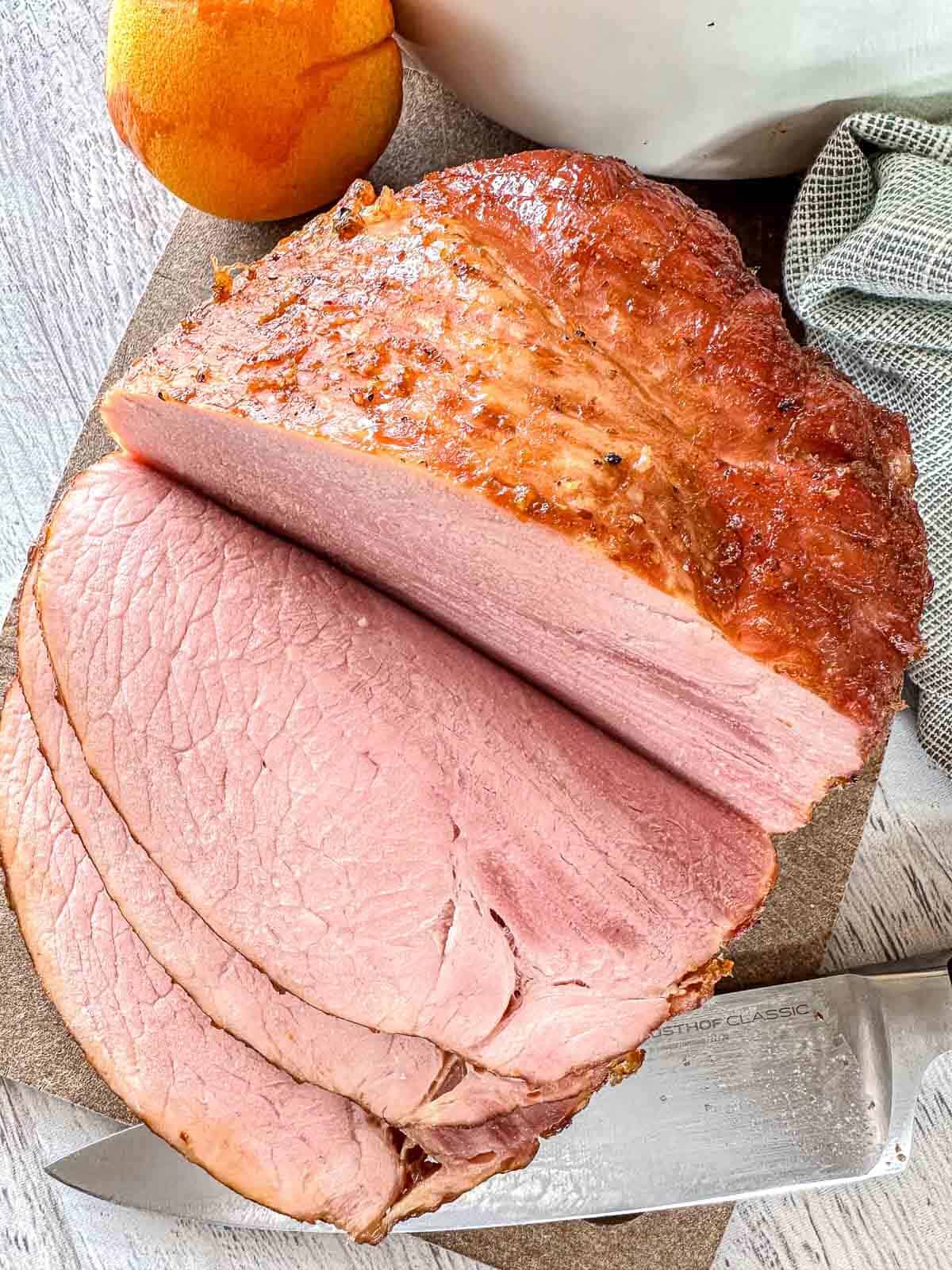 A sliced baked ham with a golden-brown crust sits on a cutting board next to a knife, an orange, and a textured cloth. The ham is moist and pink inside, showing several thick, juicy slices.