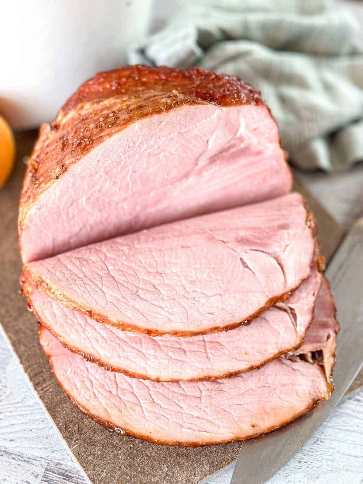 Close-up of a cooked Dutch Oven ham partially sliced on a cutting board with a knife nearby, showing three thick, pink slices and a textured brown crust. A green cloth is in the background.