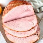 A close-up of a cooked ham, partially sliced to show its juicy, pink interior and slightly browned, caramelized outer layer. The ham sits on a cutting board with a textured cloth in the background.