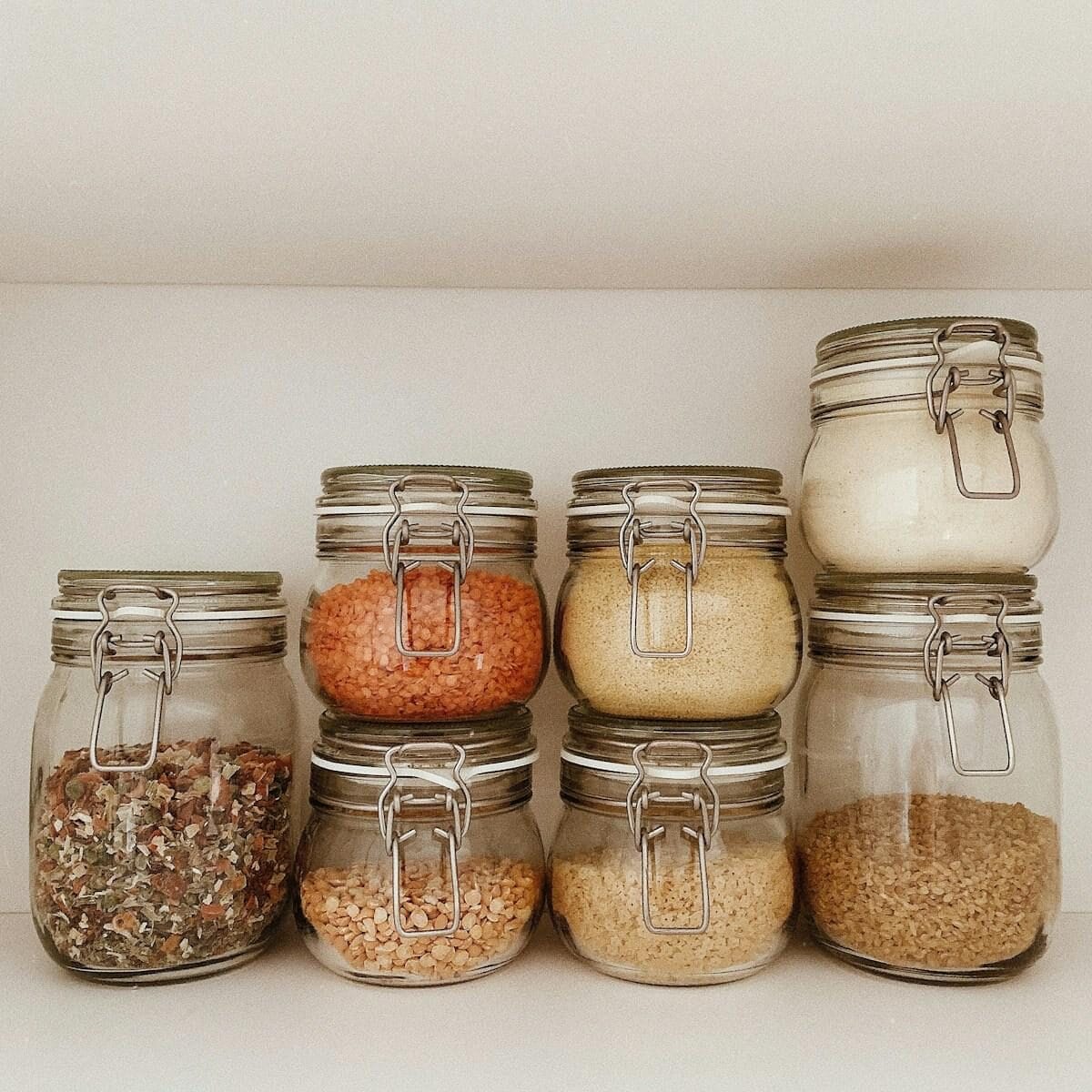 Seven glass jars with metal clasps, filled with various dry food items such as grains, lentils, and powders, are neatly arranged on a white shelf.