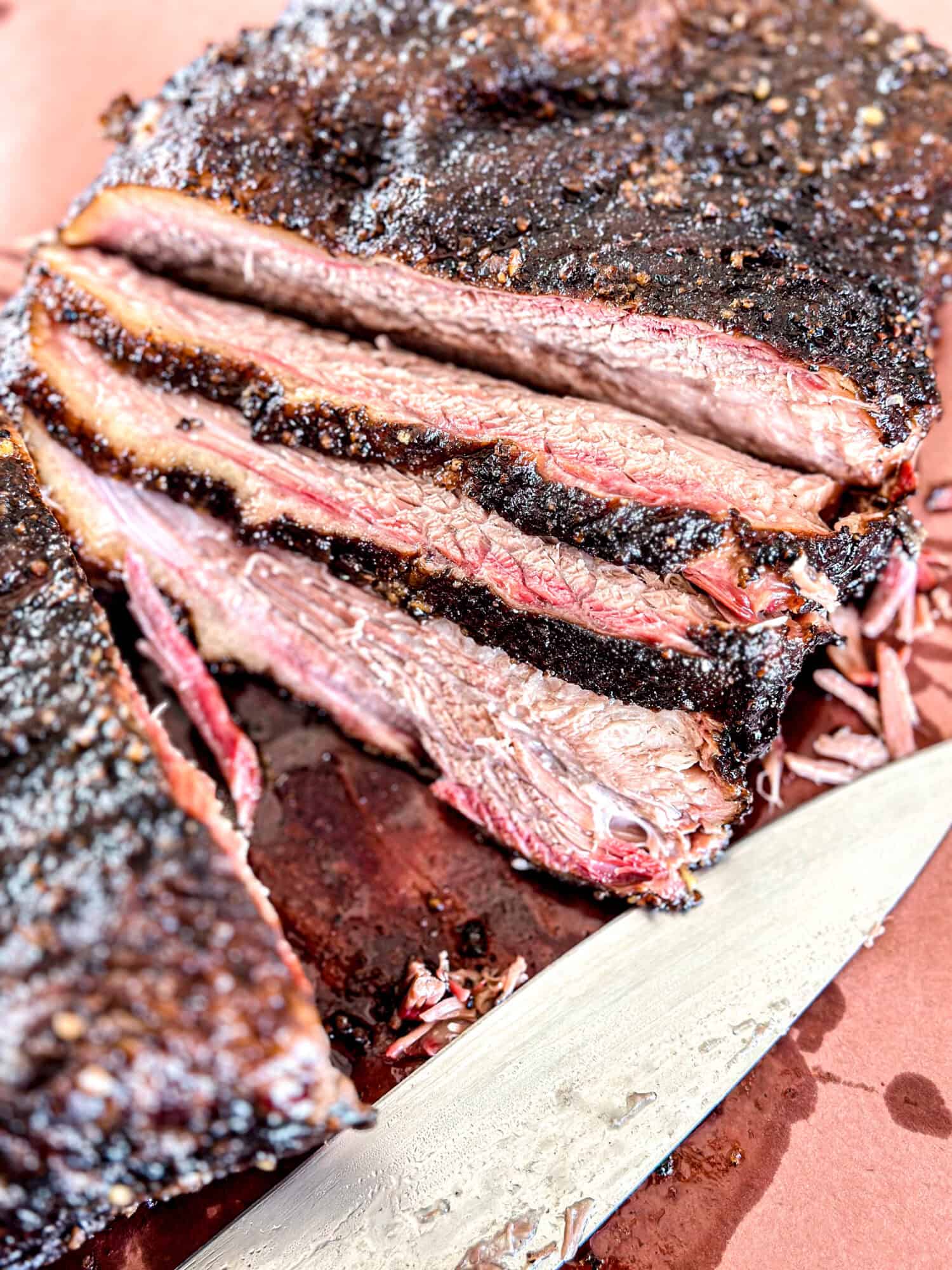 Sliced smoked brisket with a dark, seasoned crust and a visible smoke ring, arranged on a pink surface next to a large knife. Juicy meat and texture are clearly visible.