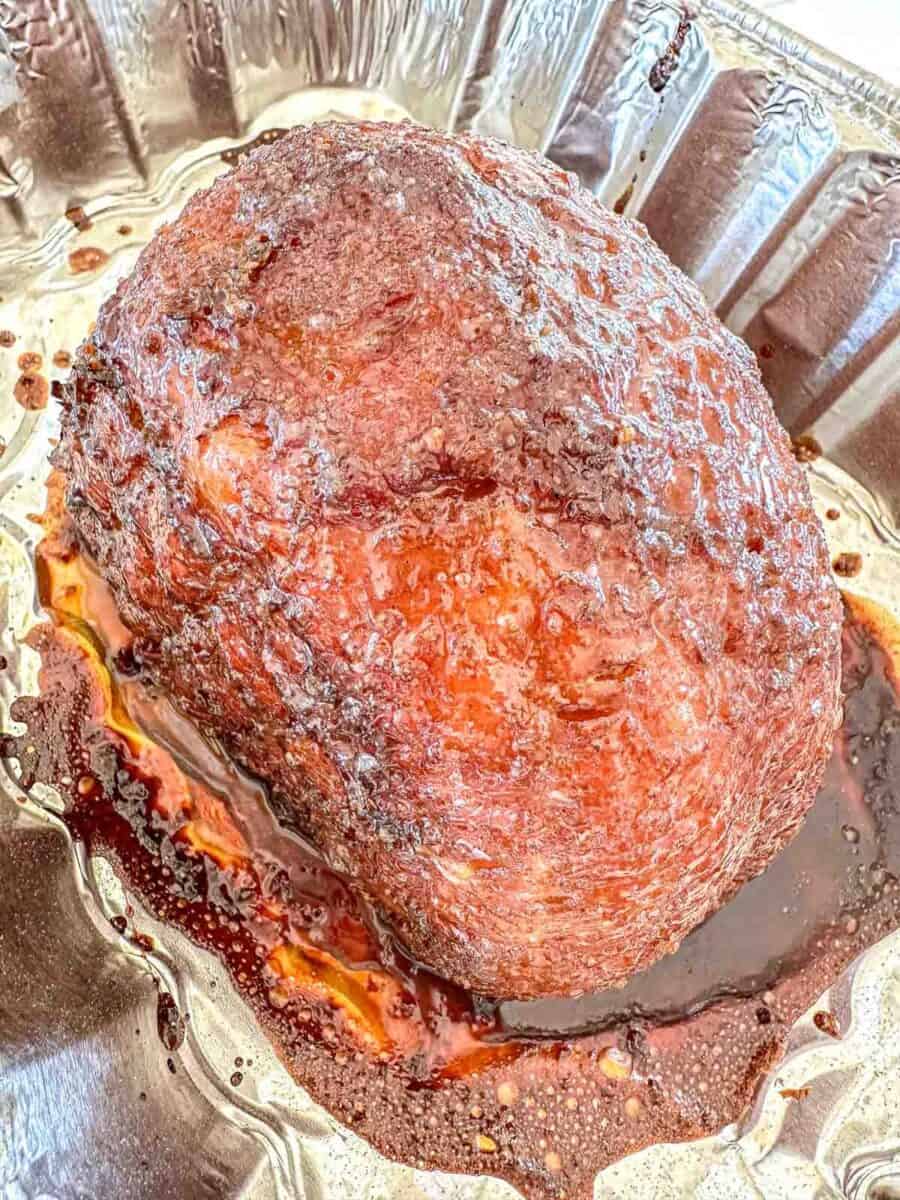 A cooked ham with a shiny, caramelized glaze sits in a disposable aluminum tray, surrounded by juices and glaze drippings.