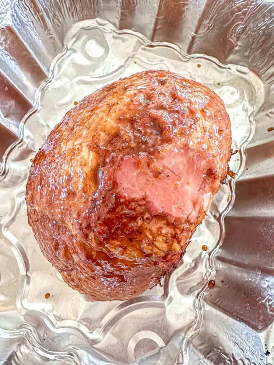 A cooked ham with a glazed, browned surface sits in a silver, scalloped roasting pan.