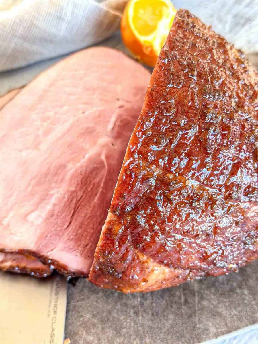 A close-up of a sliced baked ham with a caramelized, crispy outer layer. The tender, pink interior contrasts with the textured, brown crust. An orange half is visible in the background.