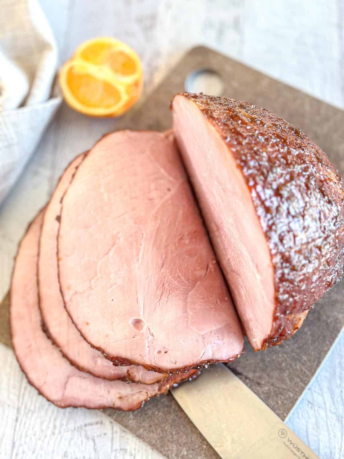 A partially sliced smoked ham on a cutting board with a knife underneath, next to a halved lemon in the background. The ham has a dark, seasoned crust.