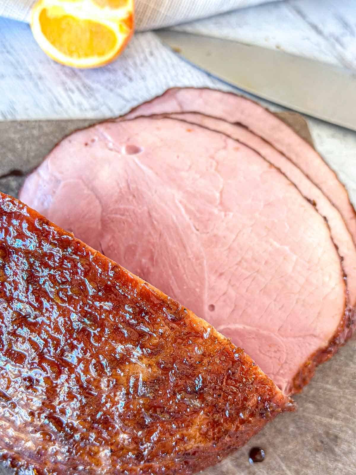 Sliced glazed ham on a cutting board with a knife nearby and an orange slice in the background. The ham has a browned, caramelized crust and a pink, juicy interior.