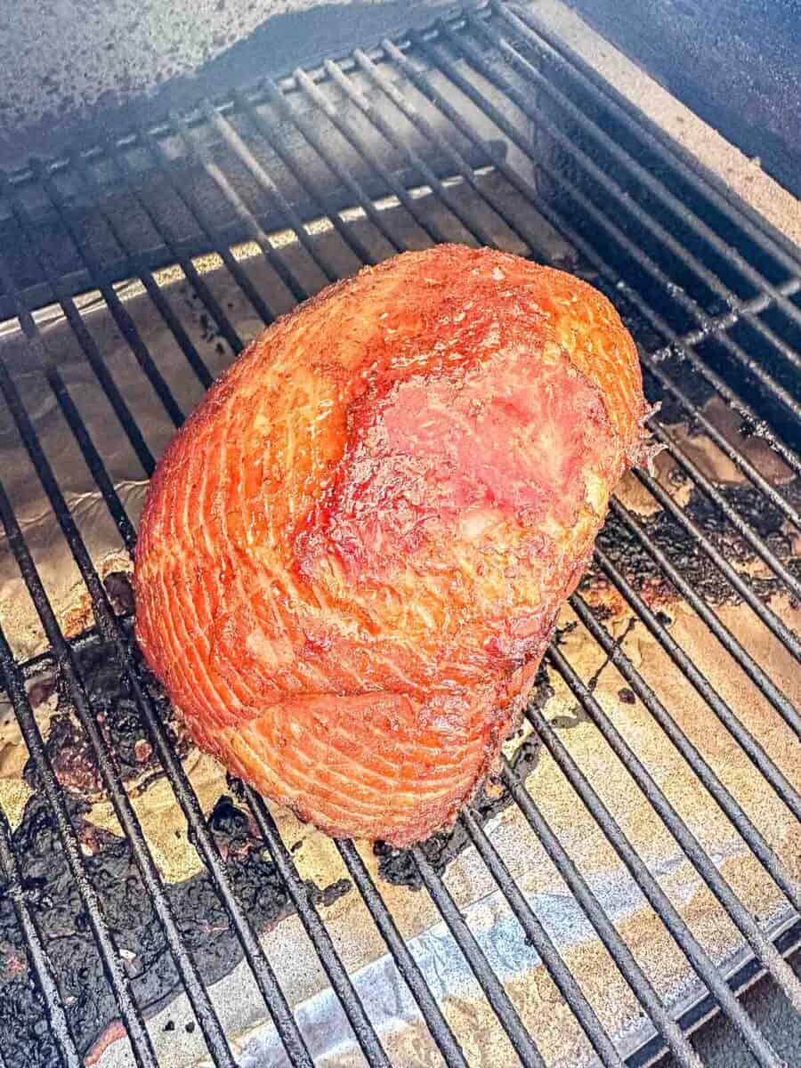 A smoked ham with a caramelized, reddish-brown glaze sits on the grates of a grill or smoker, with visible grill marks and a slightly charred surface.