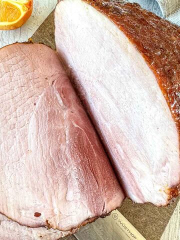 A close-up of a sliced, smoked ham on a cutting board, with a portion of an orange visible in the top left corner. The ham has a browned crust and pink, juicy interior.
