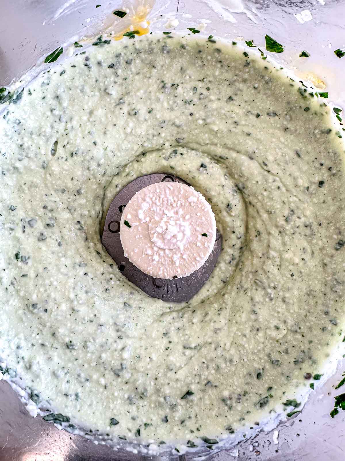 Close-up of a food processor bowl filled with a creamy, pale green mixture with visible green herb flecks. The metal blade is partially visible in the center.