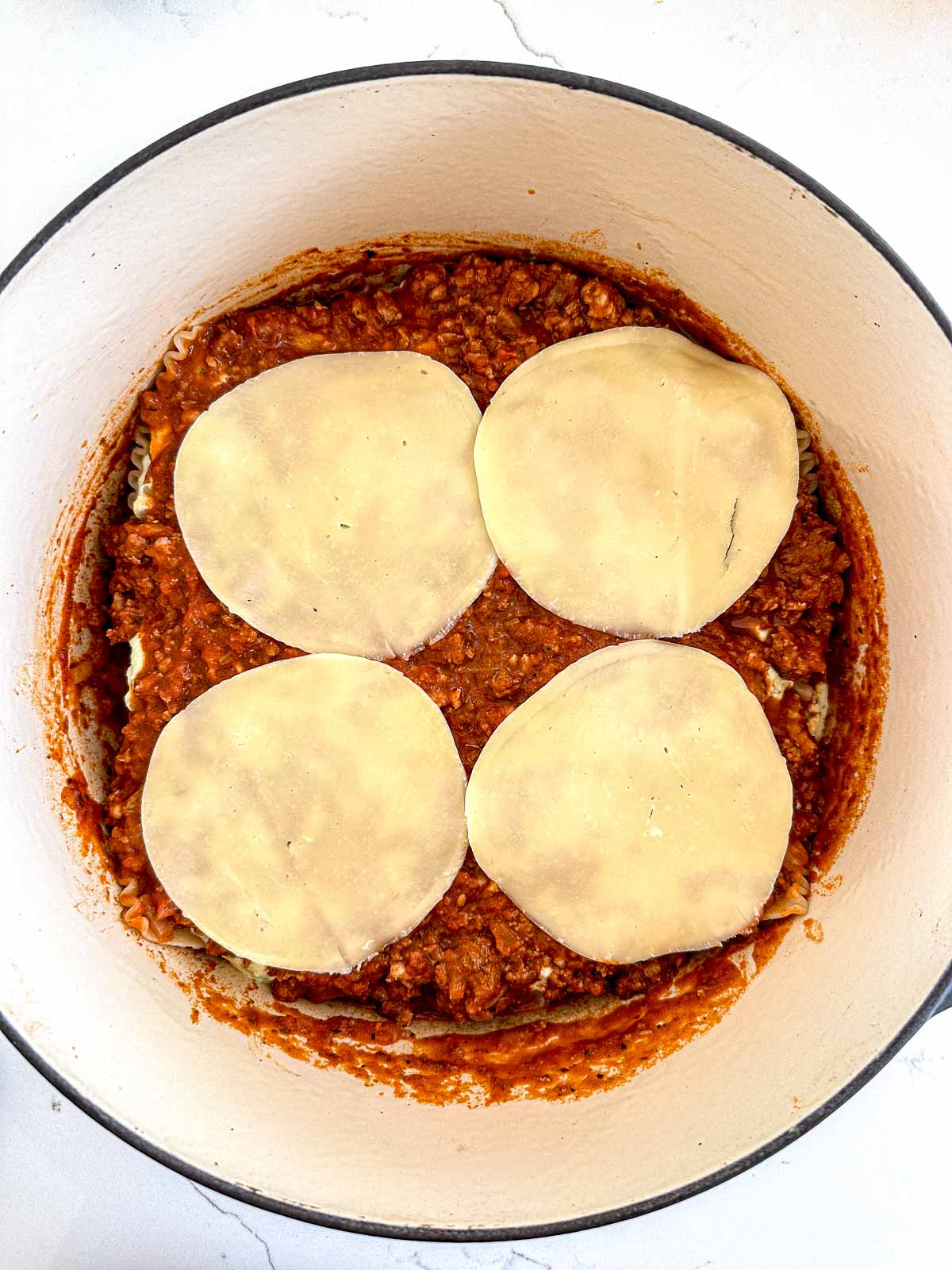 A white pot filled with a layer of meat sauce topped with four round slices of cheese, part of a lasagna preparation process.