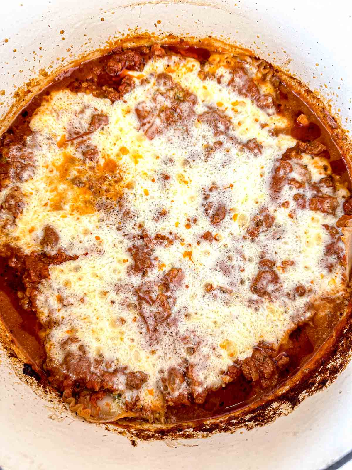 A close-up of a Dutch oven filled with bubbling, cheesy, baked lasagna, featuring browned ground meat, beans, tomato sauce, and melted cheese on top. The sides of the pot are stained with baked sauce.