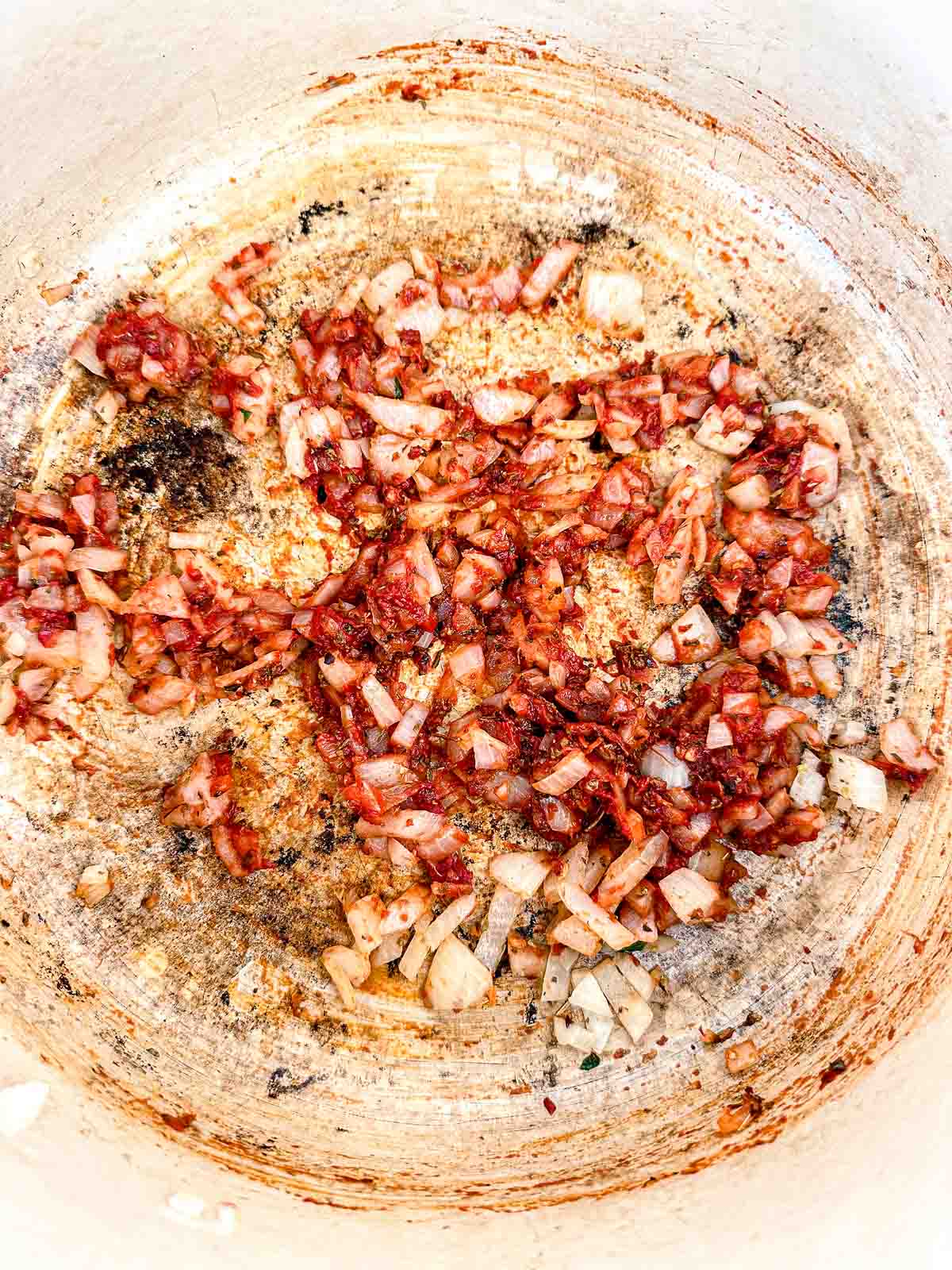 Chopped onions sautéing in a white pan, turning golden brown with bits of seasoning and browned bits sticking to the pan surface.