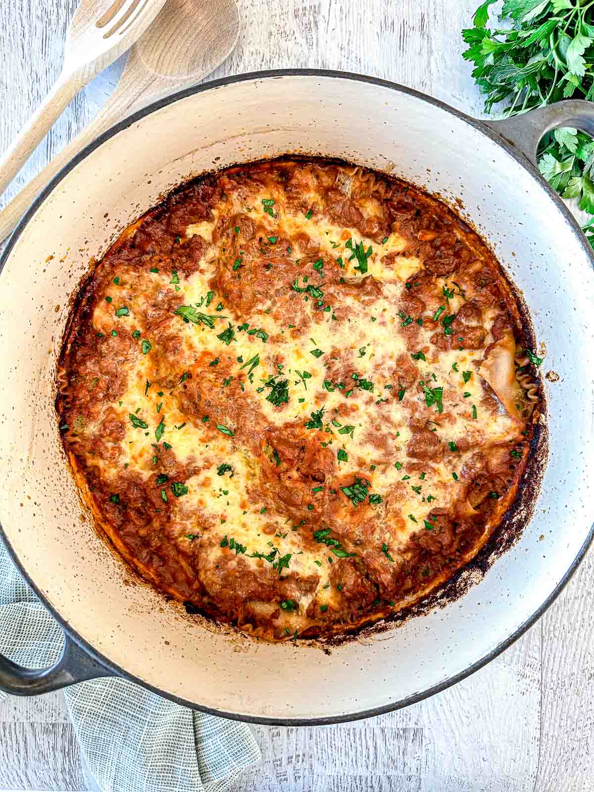 A baked lasagna with a golden, bubbly cheese crust, garnished with chopped parsley, in a white Dutch oven on a light wooden surface with a wooden spoon, fork, and fresh herbs nearby.