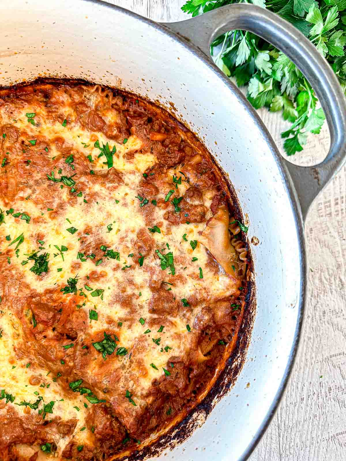 A baked lasagna with a golden, cheesy top and sprinkled parsley, in a white Dutch oven, sits on a light wooden surface with fresh parsley nearby.