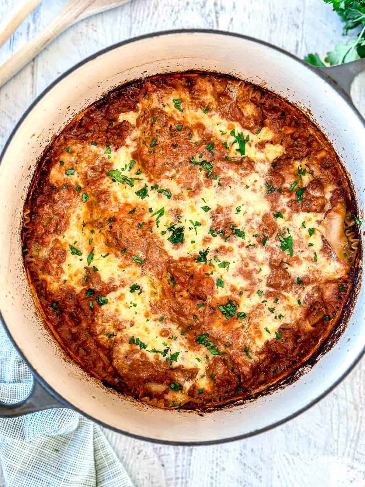 A baked lasagna with a golden, bubbly cheese topping and sprinkled with chopped parsley, shown in a round, white Dutch oven on a light wooden surface.