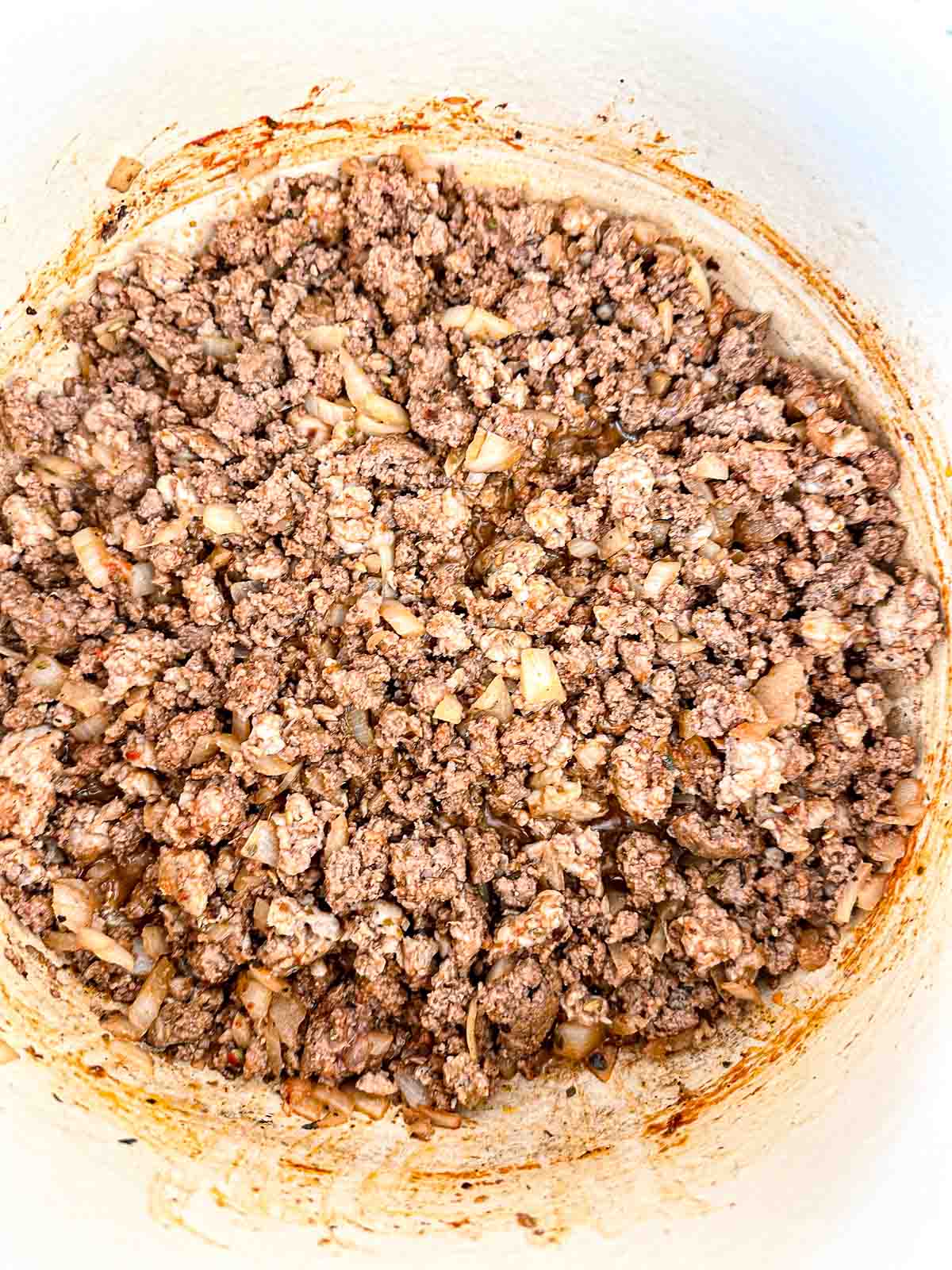 Ground beef and sausage cooked with chopped onions in a white pot, showing a mix of browned meat and translucent onions with some seasoning visible on the sides of the pot.
