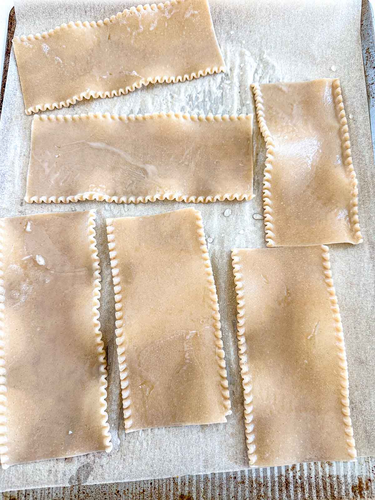 Six uncooked lasagna noodles with ruffled edges are laid out on a sheet of parchment paper, ready for use.