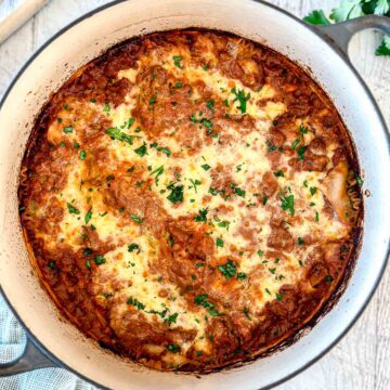 A Dutch oven lasagna with a golden, bubbly cheese topping, garnished with chopped parsley, in a white round casserole dish on a light wooden surface.