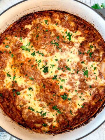A Dutch oven lasagna with a golden, bubbly cheese topping, garnished with chopped parsley, in a white round casserole dish on a light wooden surface.