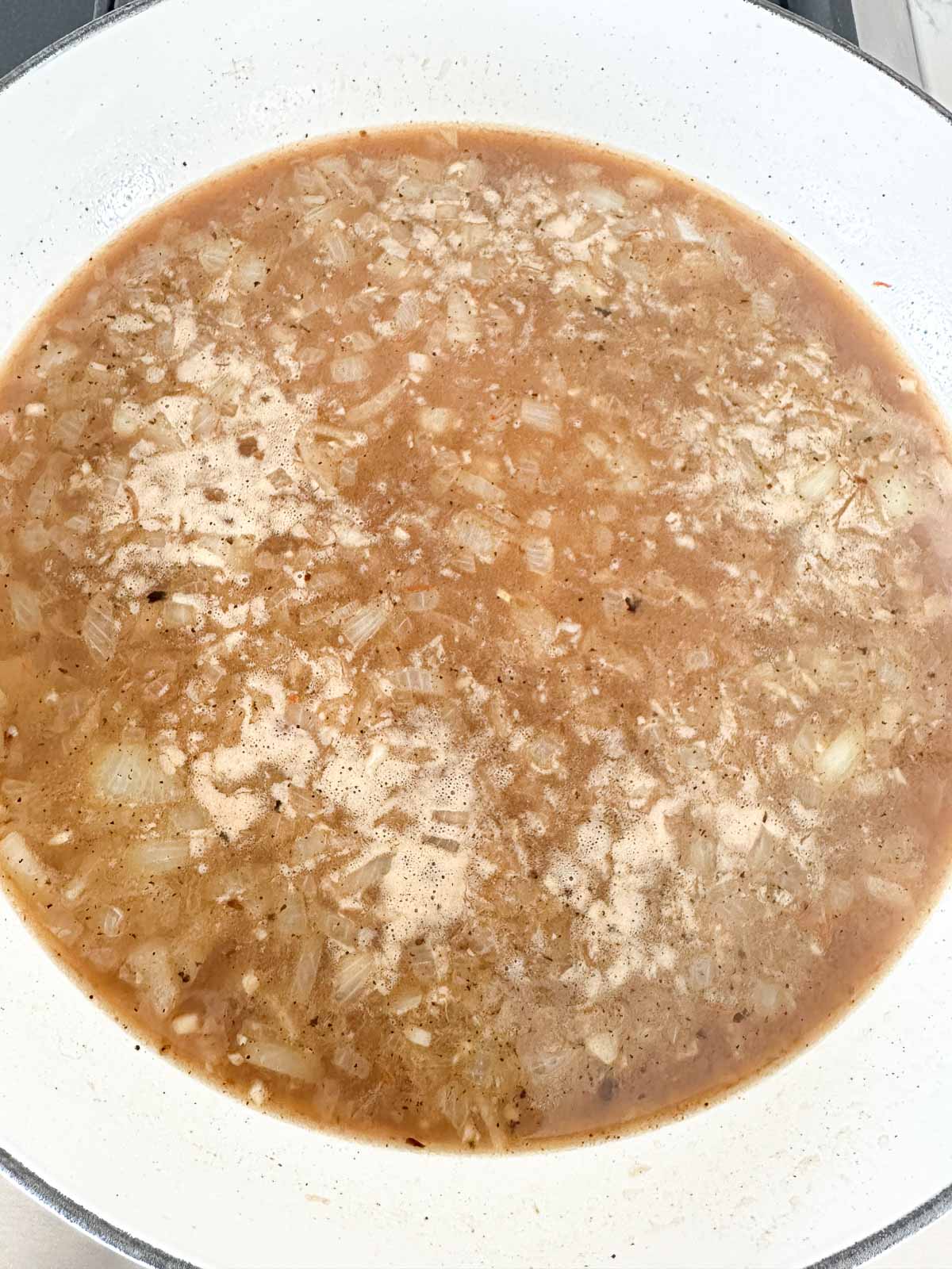 A close-up of a white pan filled with a simmering mixture of diced onions, seasonings, and broth, showing a light brown, slightly cloudy liquid with small bits floating on the surface.