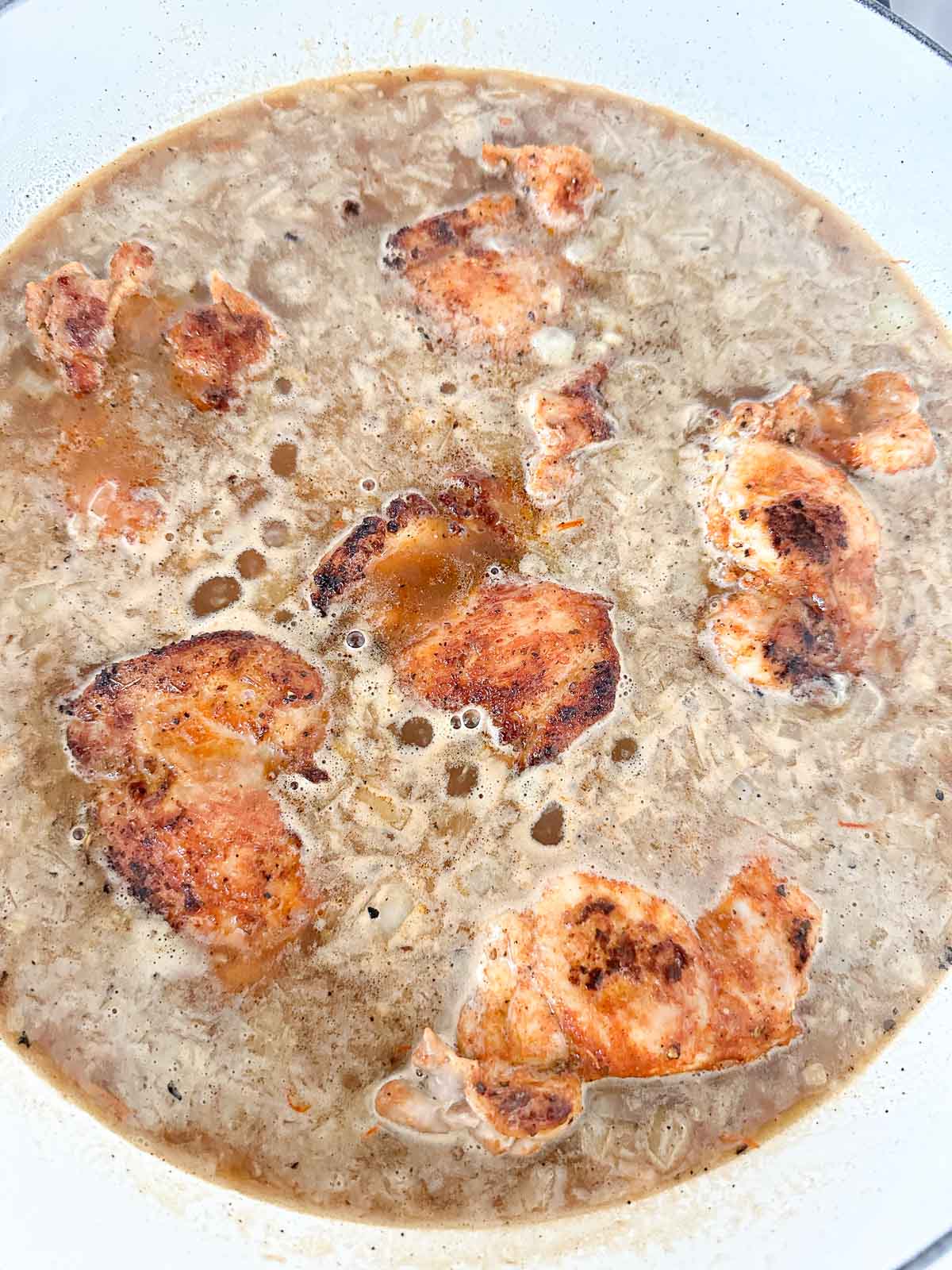 Chicken pieces simmer in a pot of brown, bubbling broth with visible bits of onion and spices. The chicken appears browned and partially submerged in the liquid.