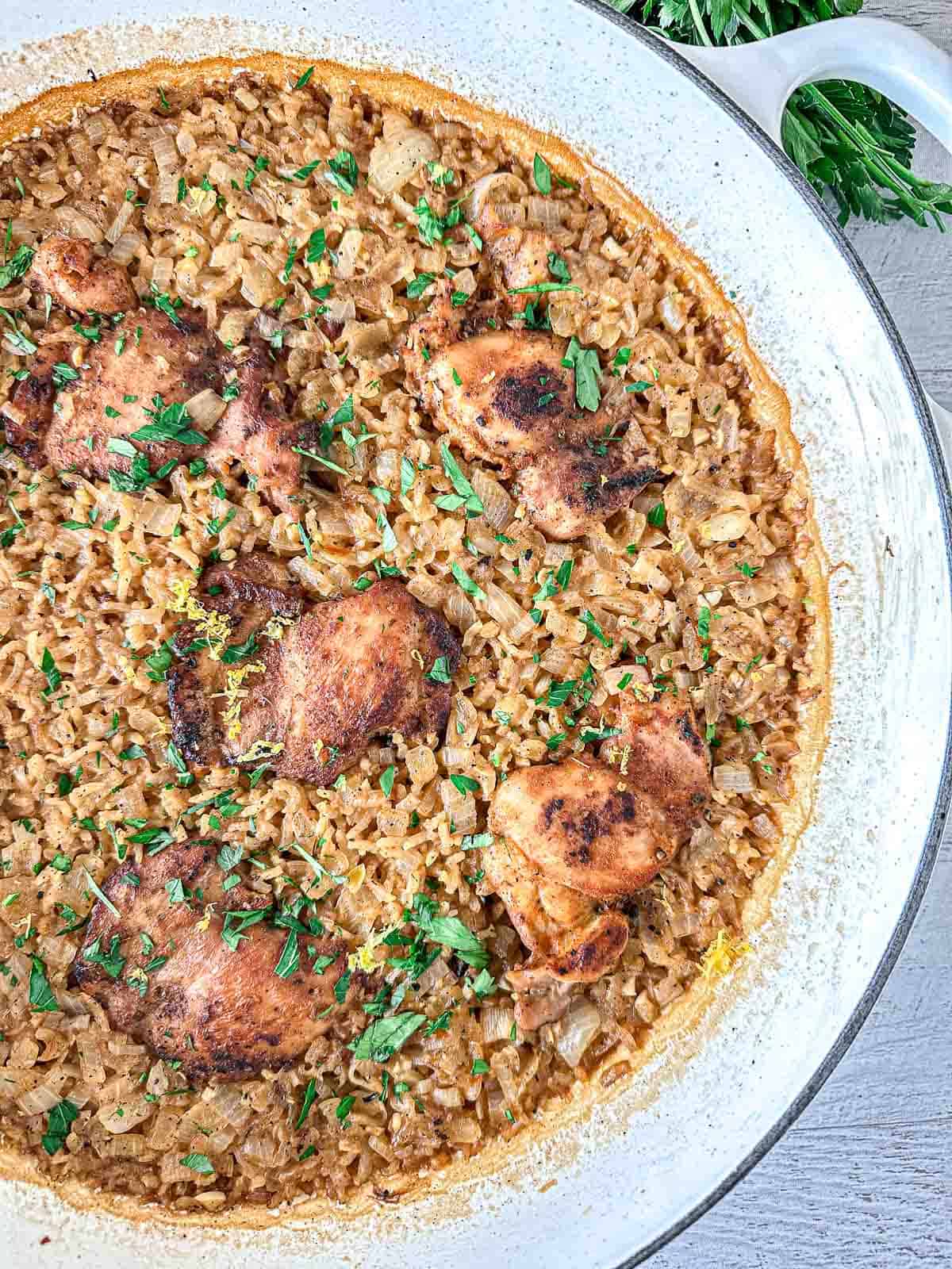 A skillet filled with baked chicken thighs on top of seasoned rice, garnished with chopped fresh herbs. The chicken is browned and the rice appears fluffy and flavorful.