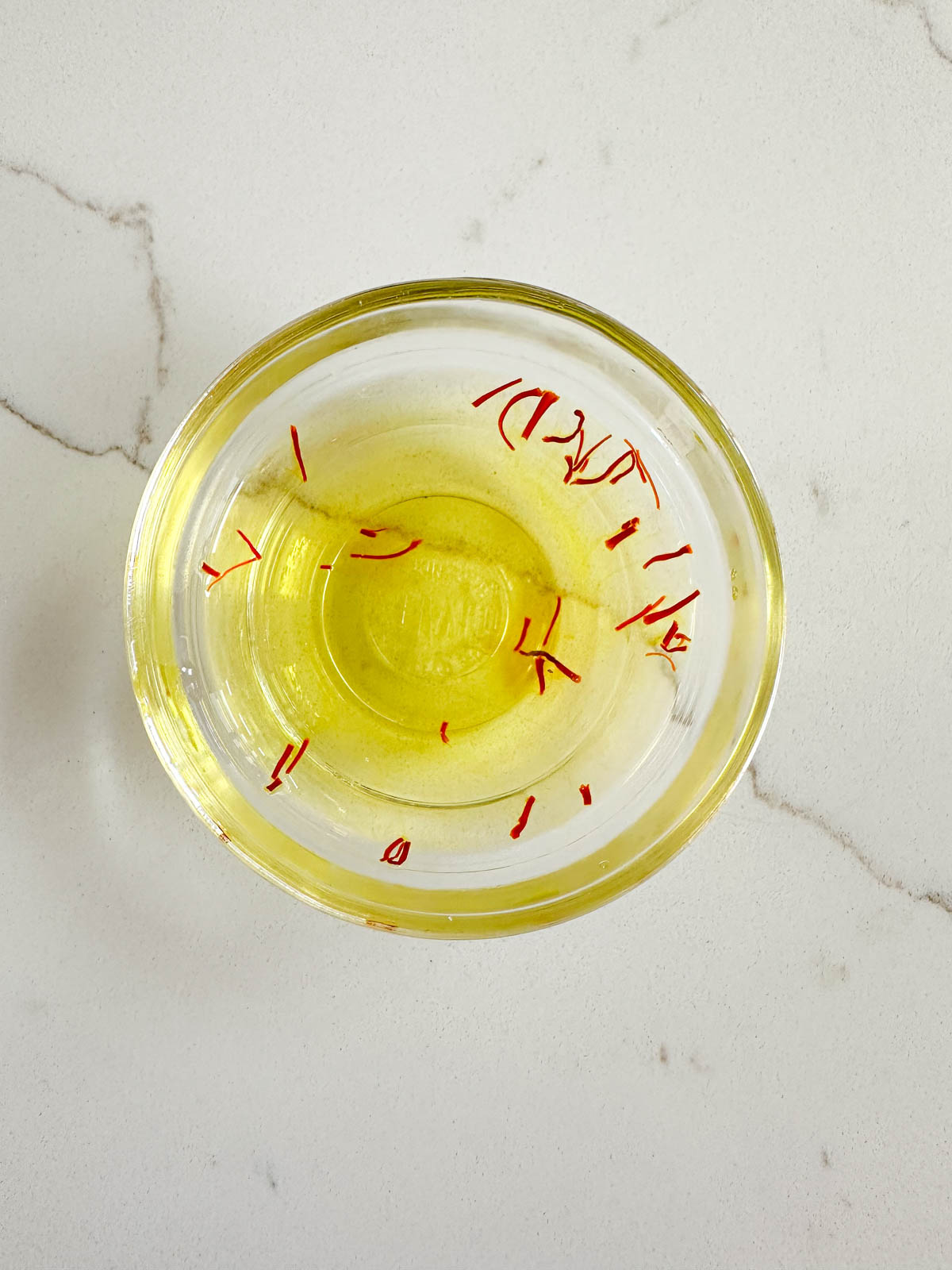 A small glass bowl filled with yellow liquid and red saffron threads, placed on a white marble surface.