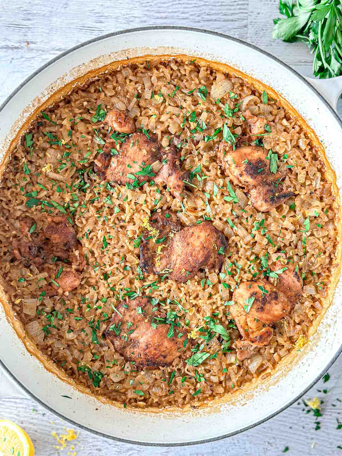 A large white skillet filled with golden-brown chicken thighs on top of seasoned rice, garnished with chopped fresh parsley. The dish rests on a light surface with herbs and lemon nearby.