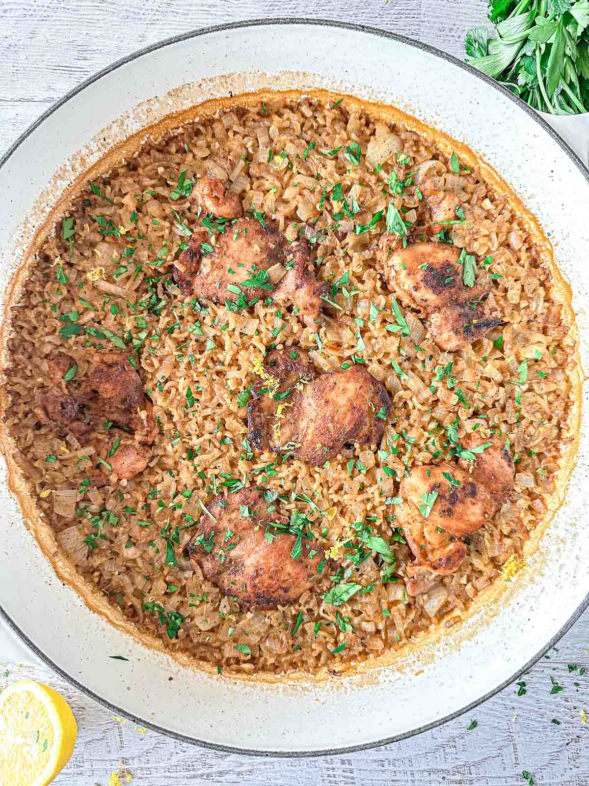 A large Dutch oven skillet filled with seasoned chicken thighs and rice, garnished with chopped fresh herbs. The dish is cooked and golden brown, with visible onions in the rice. Parsley and a lemon wedge are beside the skillet on a white surface.